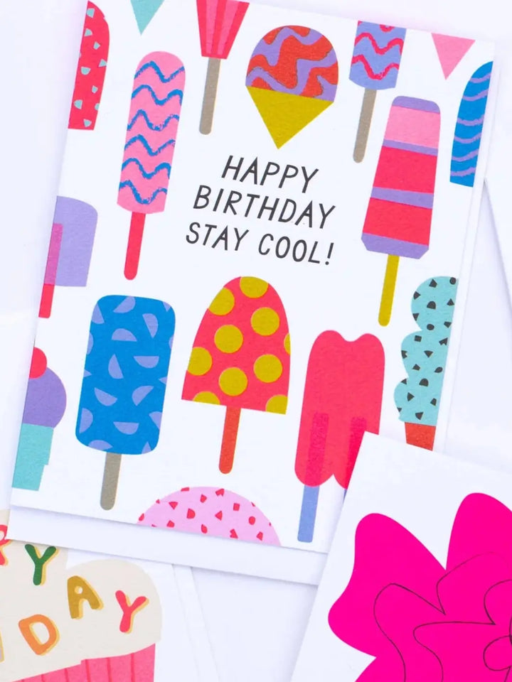 An angled, close-up shot shows the Banquet Workshop Icy Treats Happy Birthday Stay Cool Card displayed on a white surface overlapping other colorful illustrated greeting cards | Twentyseven