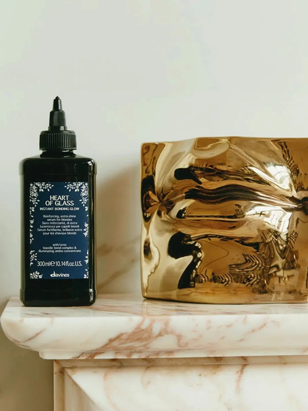 A dark bottle of Davines Heart of Glass Instant Bonding Glow stands on a marble mantelpiece next to a shiny, crumpled gold vase | Twentyseven Toronto