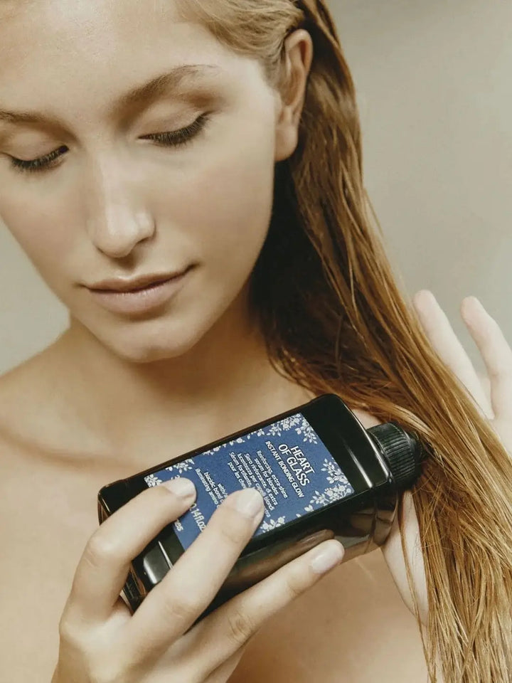 A woman with damp hair holds a bottle of Davines Heart of Glass Instant Bonding Glow while running her fingers through her blonde locks | Twentyseven Toronto