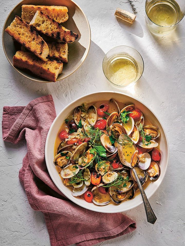 A top-down food scene showing a large white bowl filled with cooked clams, tomatoes, olives, and herbs, alongside a plate of toasted bread, two glasses of white wine, and a pink cloth on a light textured surface  Twentyseven Toronto