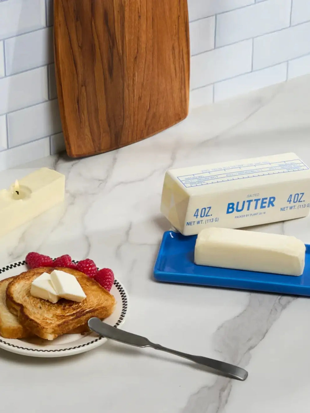 A lifestyle shot of the Item - Butter Stick Butter Dish from Fishs Eddy on a marble countertop next to a real stick of butter, toast, and fresh raspberries | Twentyseven Toronto