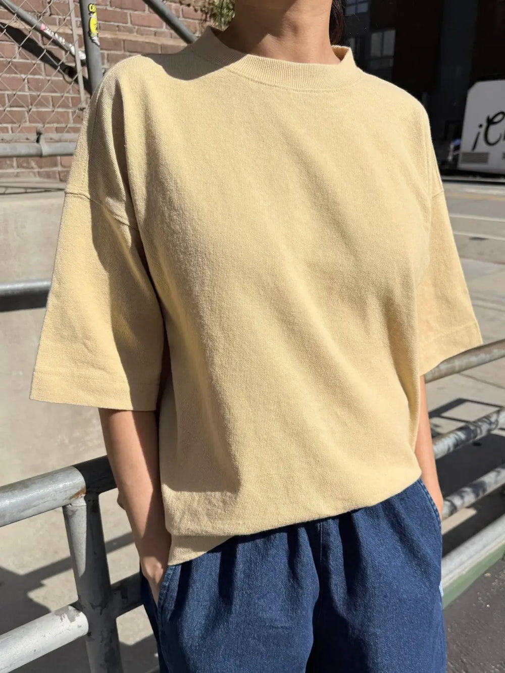 A person models the yellow oversized, short-sleeved Le Bon Shoppe Jayden Tee - Butter while standing outdoors next to a metal railing | Twentyseven Toronto