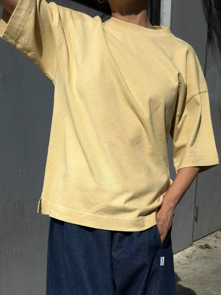 A person wearing the yellow relaxed-fit Le Bon Shoppe Jayden Tee - Butter against a gray wall, showcasing the side slit and drop-shoulder sleeve details | Twentyseven Toronto