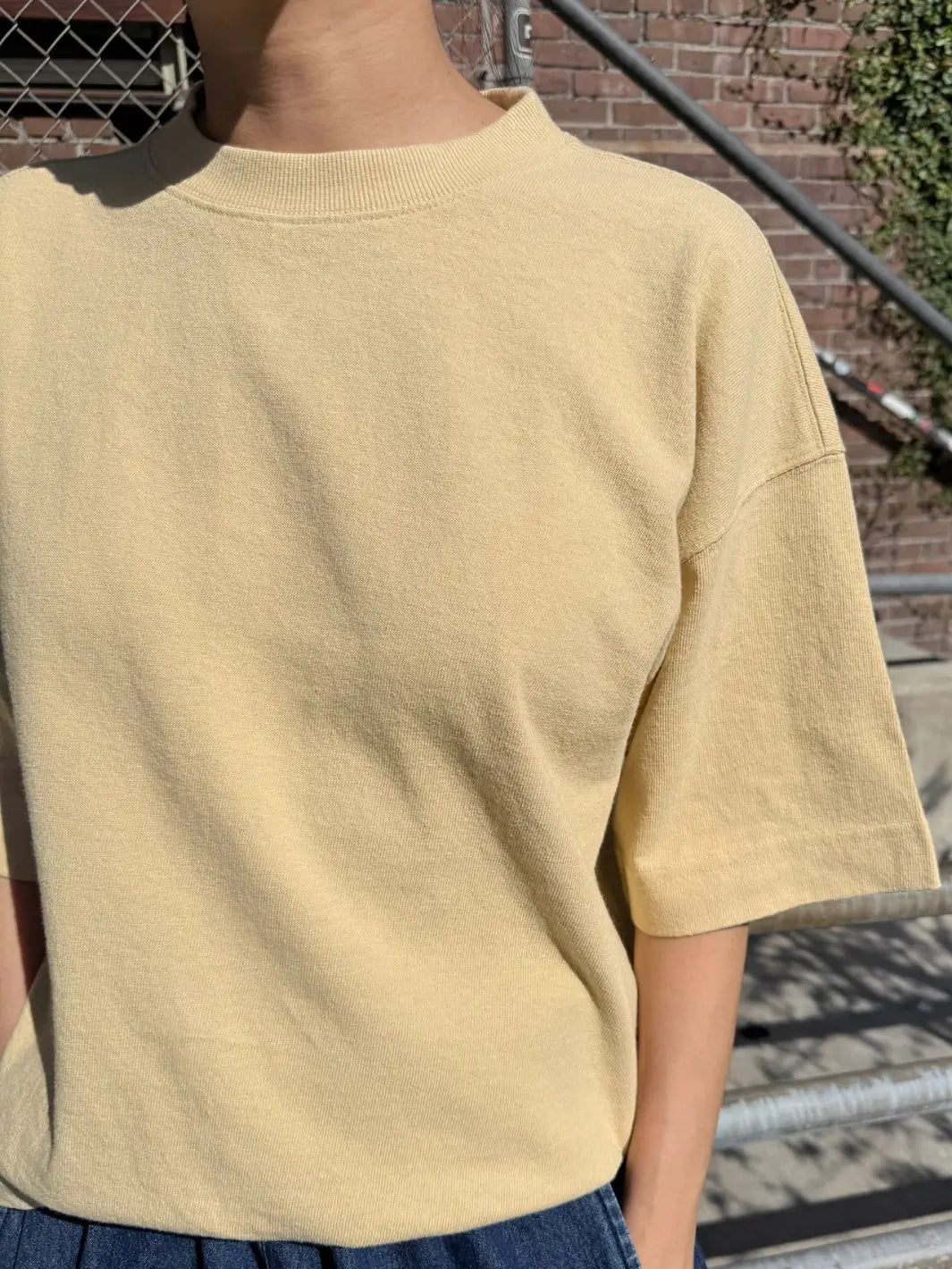 A close-up shot focuses on the textured fabric and mock neckline of the Le Bon Shoppe Jayden Tee - Butter worn by a model in an outdoor setting | Twentyseven Toronto