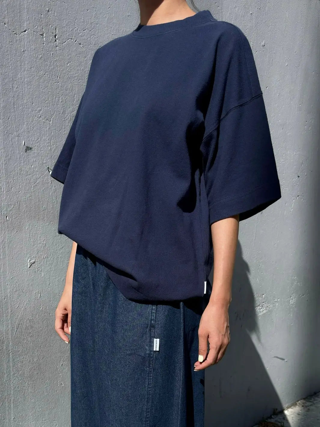 A person wears an oversized, dark navy Le Bon Shoppe Jayden Tee in Midnight paired with a denim skirt while standing against a textured gray concrete wall | Twentyseven Toronto