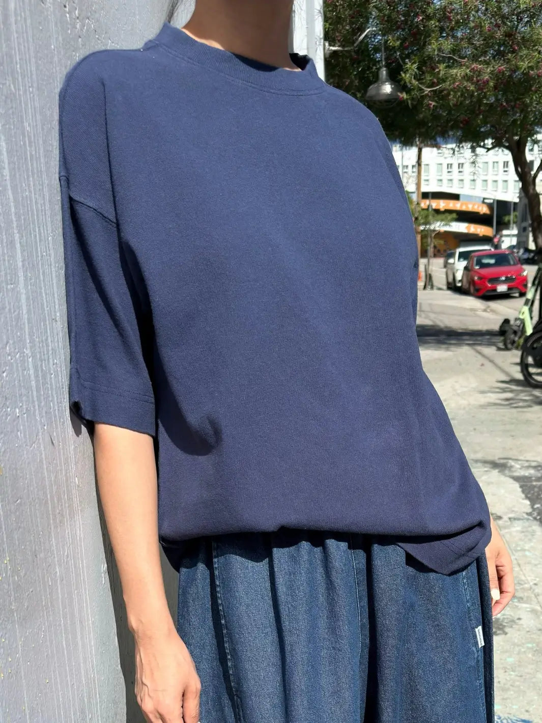 A close-up shot shows a person leaning against a light gray wall outdoors, wearing a loose-fitting Le Bon Shoppe Jayden Tee in Midnight and blue denim bottoms | Twentyseven Toronto