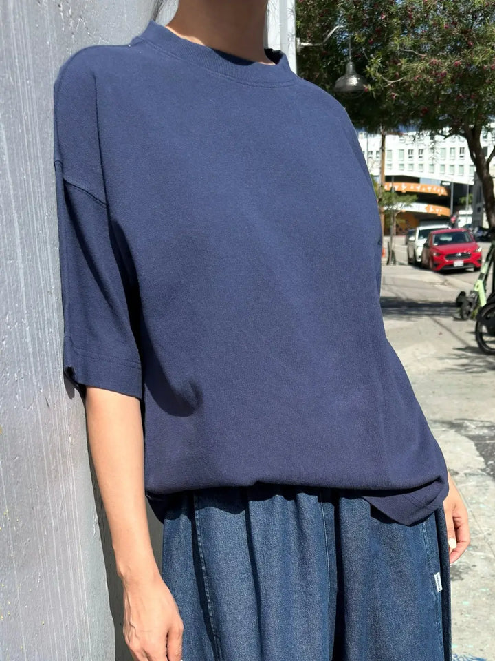 A close-up shot shows a person leaning against a light gray wall outdoors, wearing a loose-fitting Le Bon Shoppe Jayden Tee in Midnight and blue denim bottoms | Twentyseven Toronto