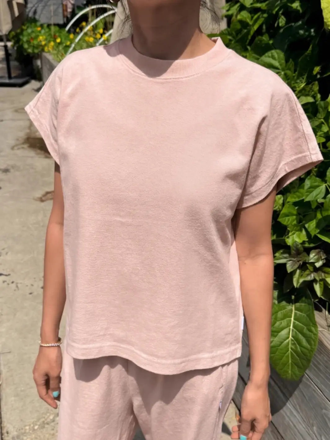A person stands outdoors wearing a boxy, short-sleeved Le Bon Shoppe Jeanne Tee in Baby Pink with matching loungewear pants against a backdrop of green plants | Twentyseven Toronto