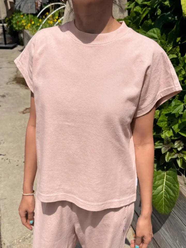 A person stands outdoors wearing a boxy, short-sleeved Le Bon Shoppe Jeanne Tee in Baby Pink with matching loungewear pants against a backdrop of green plants | Twentyseven Toronto