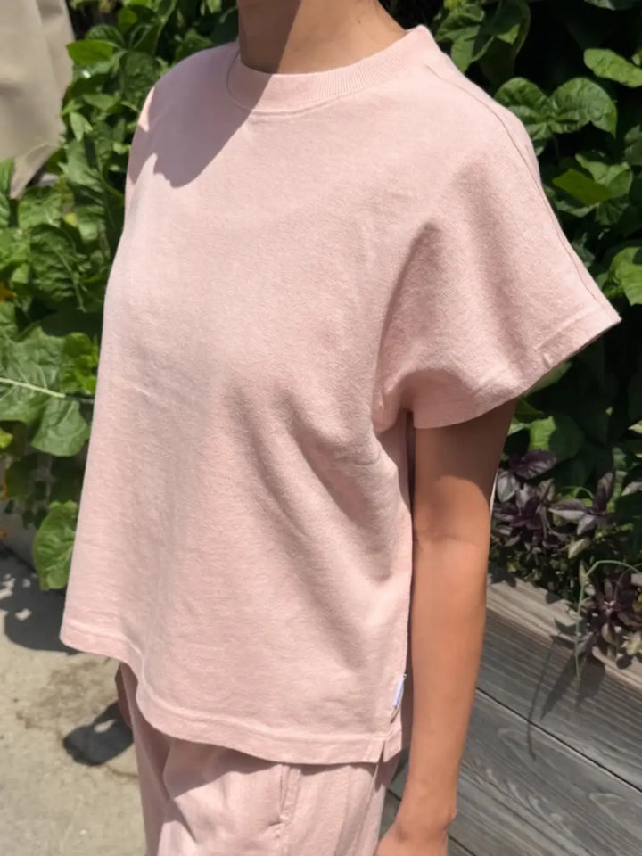 A close-up, side-angle shot shows a person wearing a light, loose-fitting Le Bon Shoppe Jeanne Tee in Baby Pink against green foliage | Twentyseven Toronto
