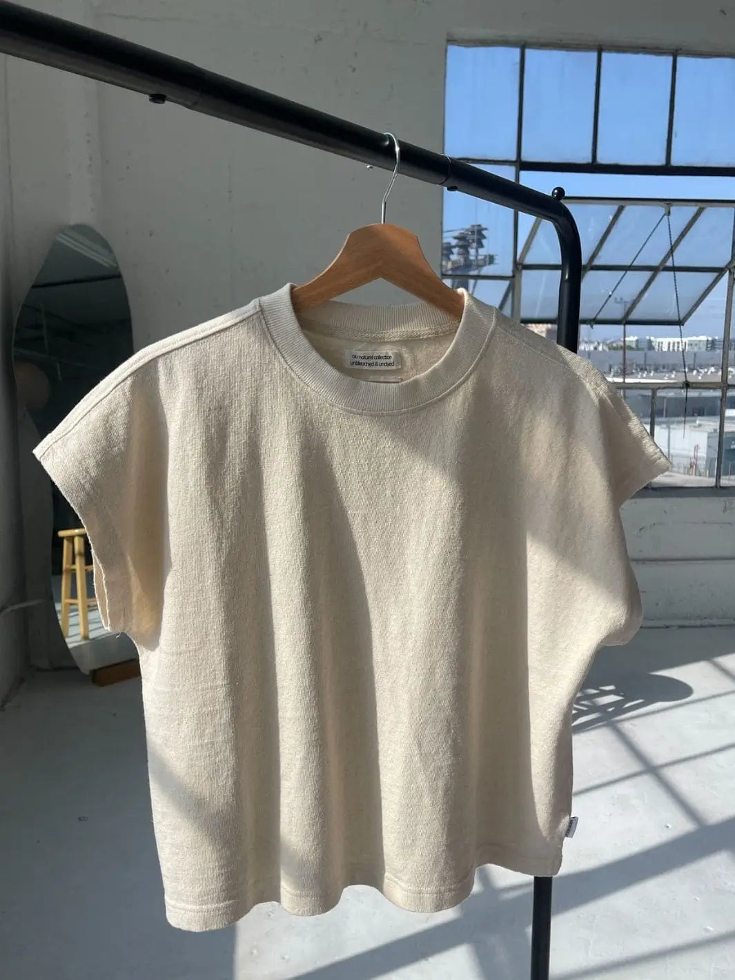 A cream-colored Le Bon Shoppe Jeanne Tee in Naturel hangs on a clothing rack inside a bright studio with large windows | Twentyseven Toronto