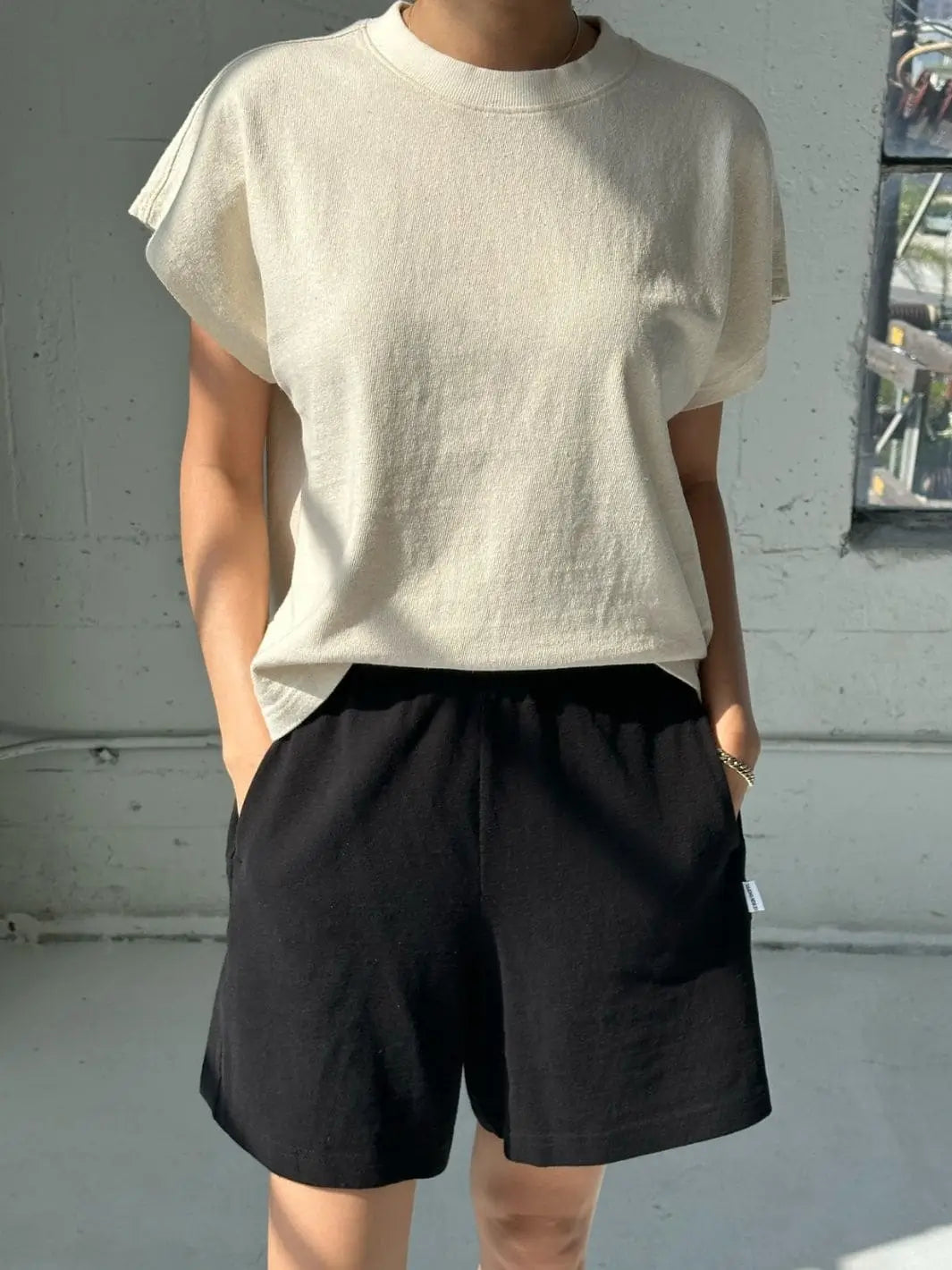 A person wears an off-white Le Bon Shoppe Jeanne Tee in Naturel tucked slightly into black drawstring shorts in a bright interior setting  | Twentyseven Toronto