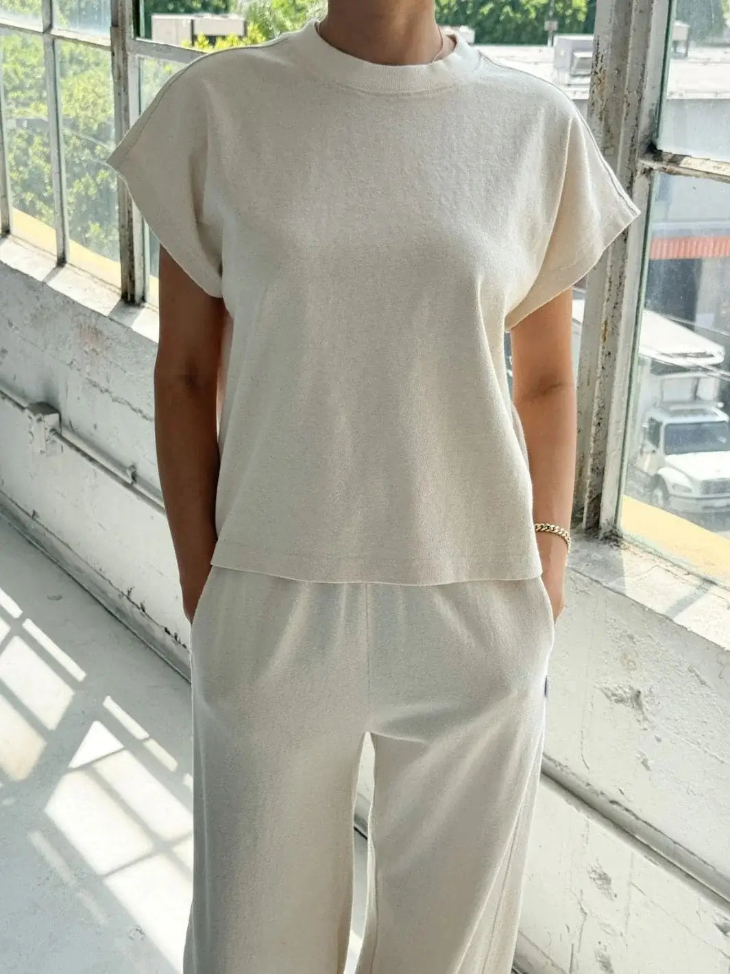A person stands near large windows wearing a matching off-white loungewear set featuring the boxy cream top from Le Bon Shoppe Jeanne Tee in Naturel | Twentyseven Toronto