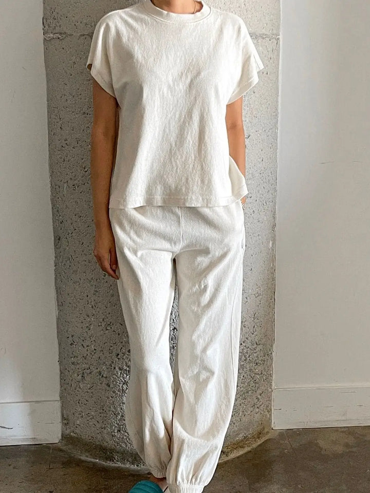 A person wears a cream-colored Le Bon Shoppe Jeanne Tee in Naturel with matching sweatpants while leaning against a textured gray concrete pillar | Twentyseven Toronto
