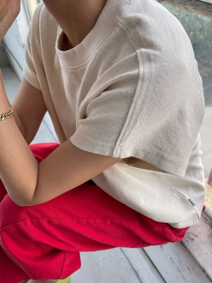 A person sits by a window wearing a textured, cream Le Bon Shoppe Jeanne Tee in Naturel and vibrant red pants | Twentyseven Toronto