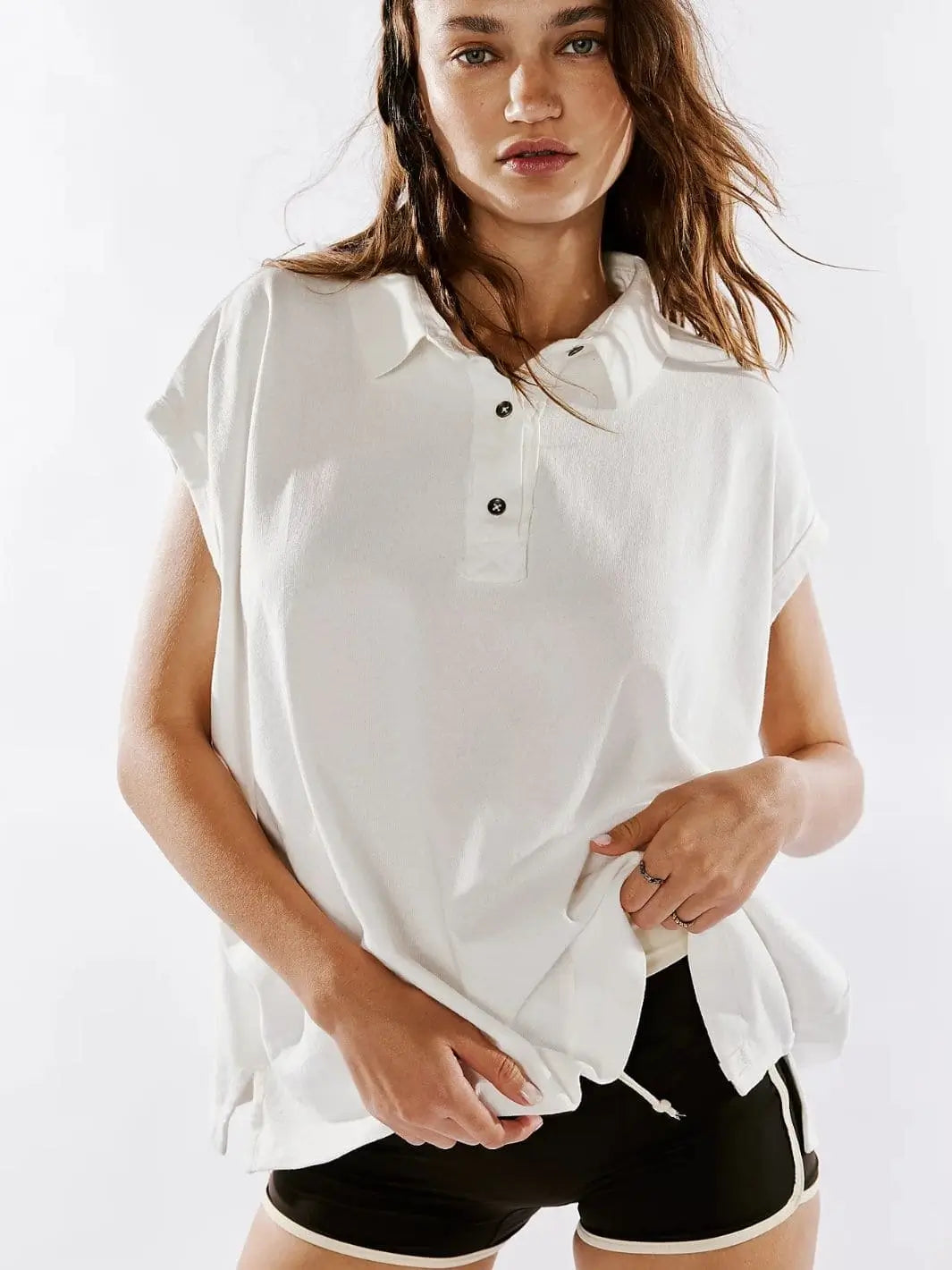 A front view shows a model with wavy hair posing in the oversized, buttoned Free People Keep On Polo - Clean Ivory paired with black shorts | Twentyseven Toronto