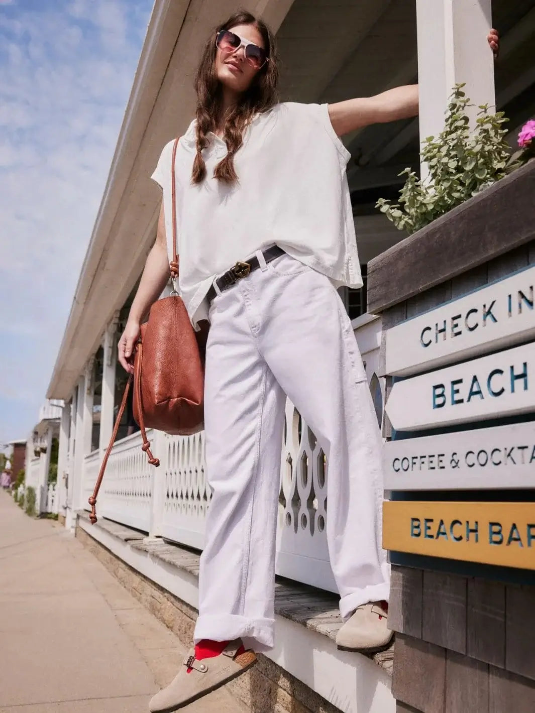 An outdoor low-angle shot shows a model wearing the Free People Keep On Polo - Clean Ivory styled with wide-leg white pants, sunglasses, and a brown leather tote bag | Twentyseven Toronto