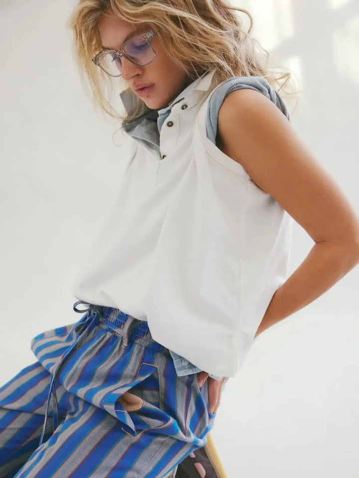 A model wearing glasses sits to show the Free People Keep On Polo - Clean Ivory layered over a blue button-down shirt and paired with blue striped pants | Twentyseven Toronto