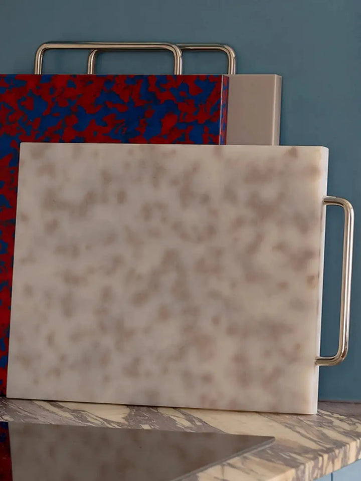 A Fredericks and Mae Large Gray/White Cutting Board with a speckled pattern and a silver metal handle, upright on a marble countertop alongside other colorful boards | Twentyseven Toronto