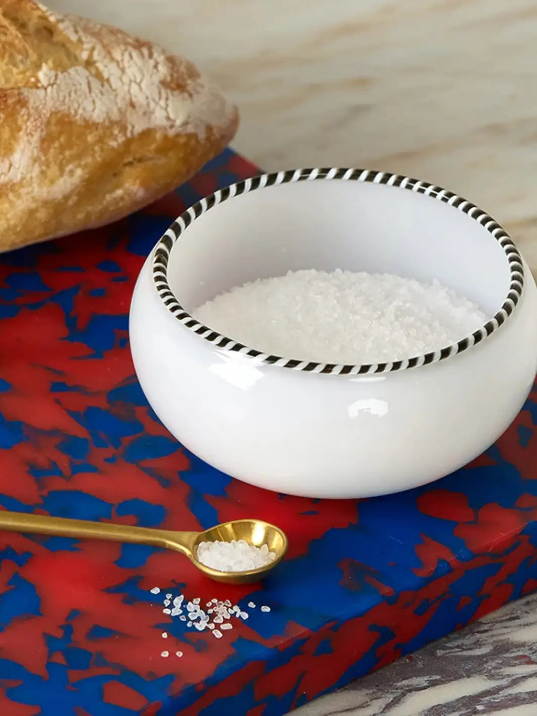 A close-up shot of a white bowl filled with coarse salt and a small gold spoon resting on a Fredericks and Mae Large Red/Blue Cutting Board next to a loaf of bread | Twentyseven Toronto