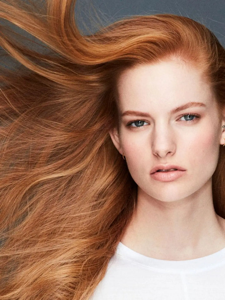 A close-up portrait of a woman with vibrant, wind-blown red hair highlights the volumizing effects of Hair by Sam McKnight Lazy Girl Dry Shampoo | Twentyseven Toronto