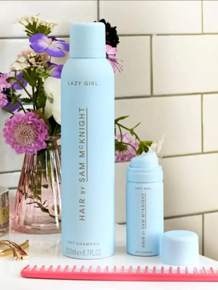 A full-size spray can and a travel-size mini version of Hair by Sam McKnight Lazy Girl Dry Shampoo are displayed on a counter next to a vase of purple flowers and a pink comb | Twentyseven Toronto