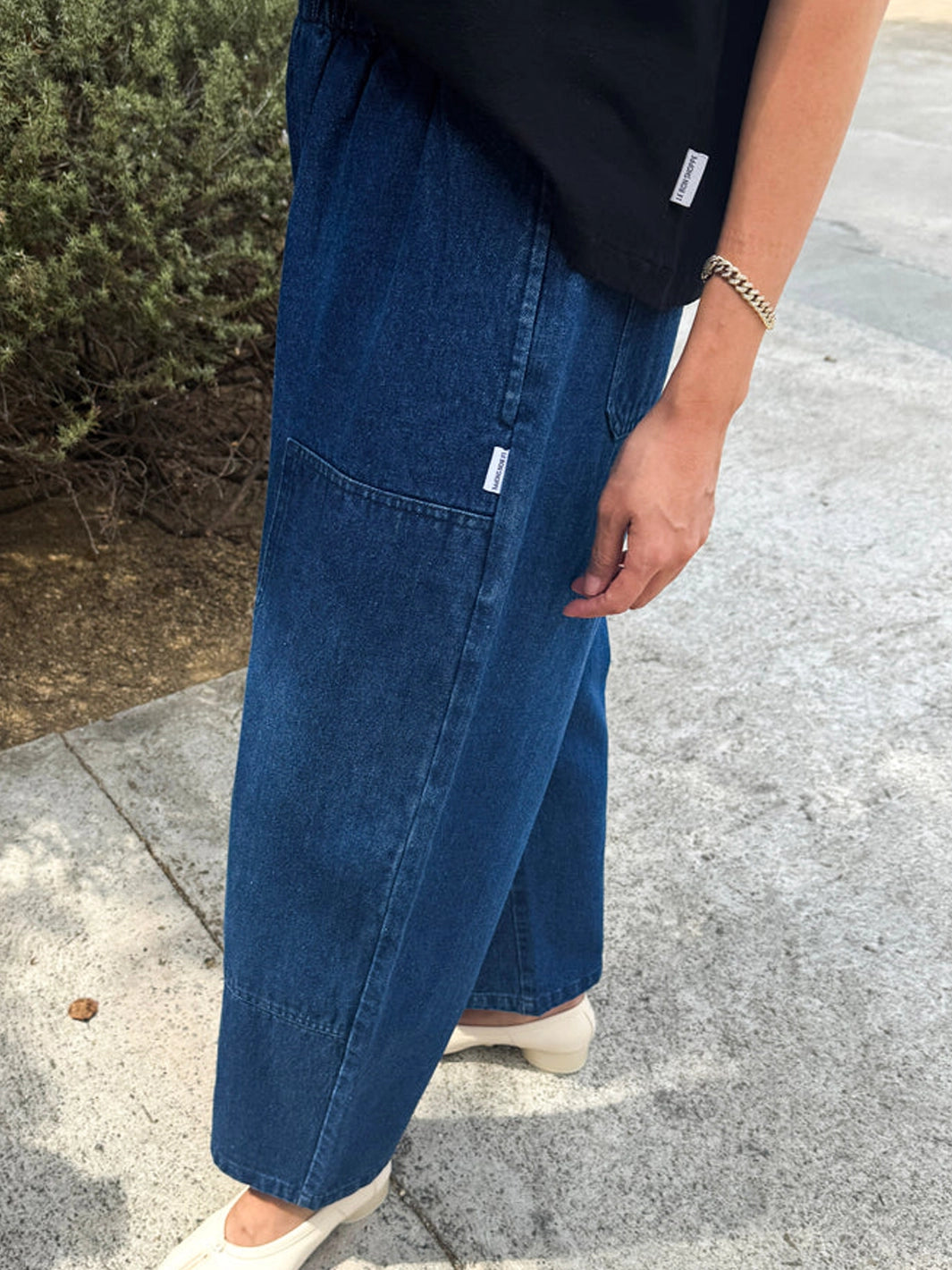 Side view of the Le Bon Shoppe Gardener’s Pants in Blue Denim styled with a black top and cream shoes, highlighting the utility-inspired patch pocket and relaxed fit | Twentyseven Toronto