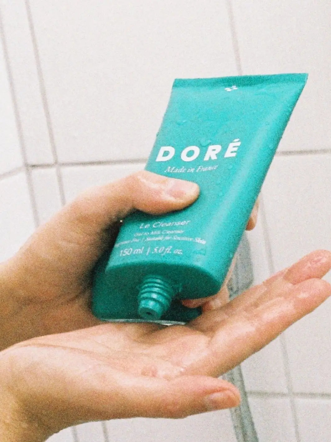 Hand squeezing a teal tube of Doré Skincare 'Le Cleanser' onto hands against a tiled wall background | Twentyseven Toronto