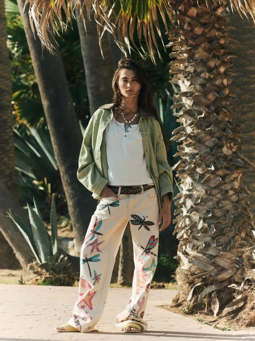 A full-length outdoor shot depicts a model standing by a palm tree wearing the Free People Lemon Denim Jacket in Washed Sage paired with illustrated white jeans | Twentyseven Toronto