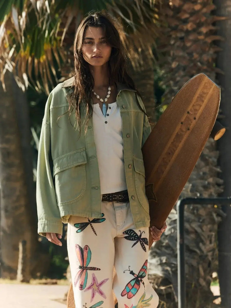 A medium outdoor shot captures the model holding a longboard while wearing the Free People Lemon Denim Jacket in Washed Sage open over a white henley top | Twentyseven Toronto