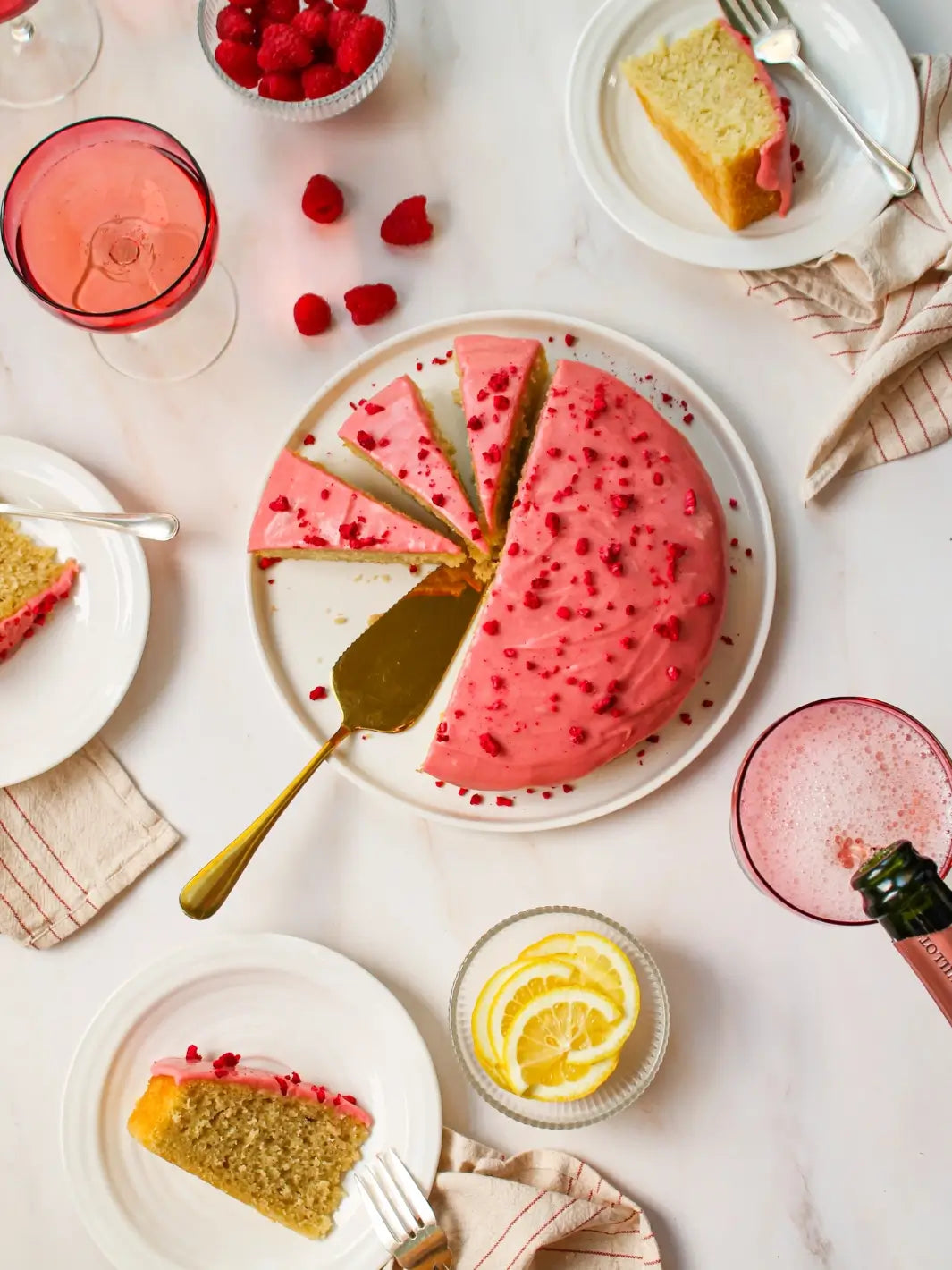A flat lay table setting features slices of cake from the Glazed Cake Co Lemon Raspberry Cake Kit served on small white plates alongside fresh raspberries, lemon slices, and sparkling wine | Twentyseven Toronto
