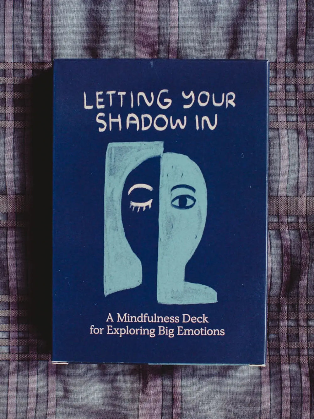 The dark blue front cover of the Letting Your Shadow In Deck from People I've Loved features a split-face illustration and sits on a plaid fabric surface | Twentyseven Toronto