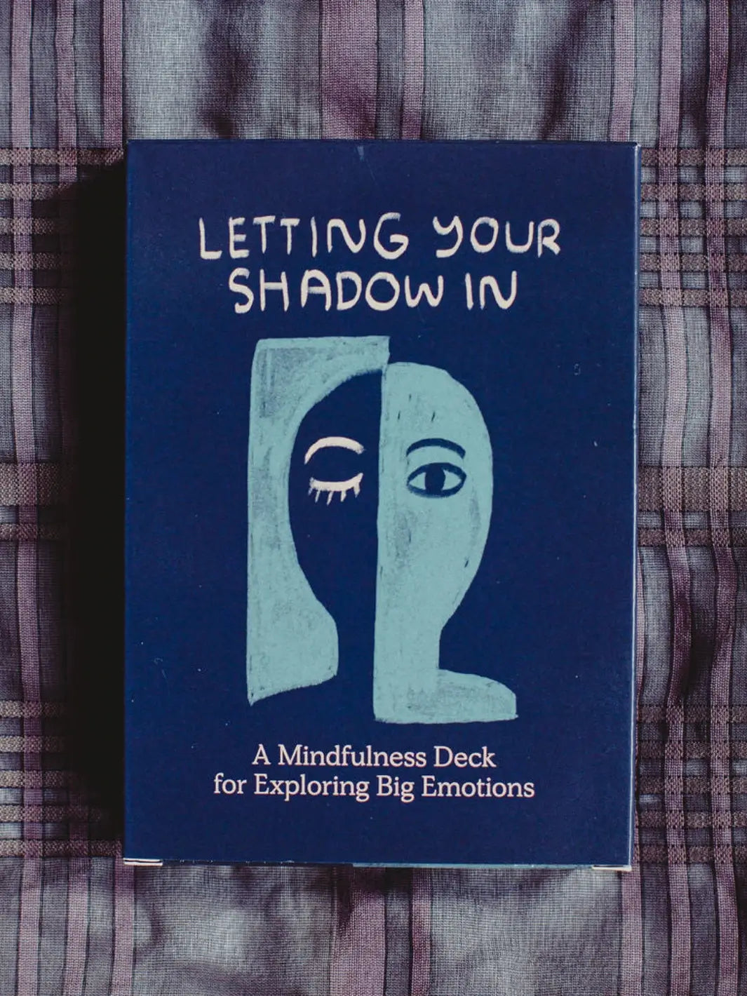 The dark blue front cover of the Letting Your Shadow In Deck from People I've Loved features a split-face illustration and sits on a plaid fabric surface | Twentyseven Toronto