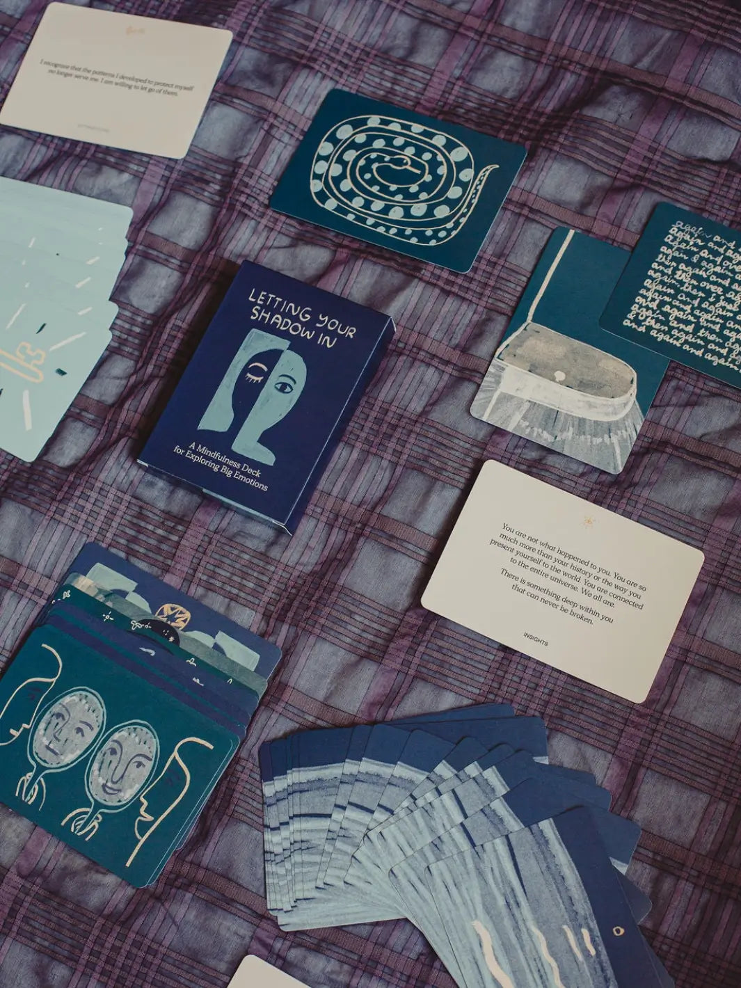 A variety of illustrated and textual cards from the Letting Your Shadow In Deck from People I've Loved are scattered across a purple plaid blanket alongside the main box | Twentyseven Toronto