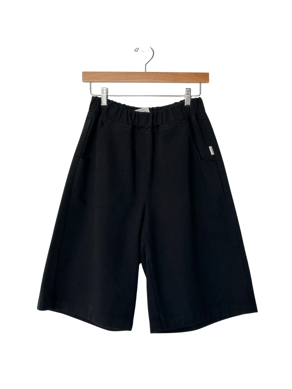 The Le Bon Shoppe Liam Bermuda Shorts - Black Canvas are displayed hanging from a wooden clip hanger | Twentyseven Toronto