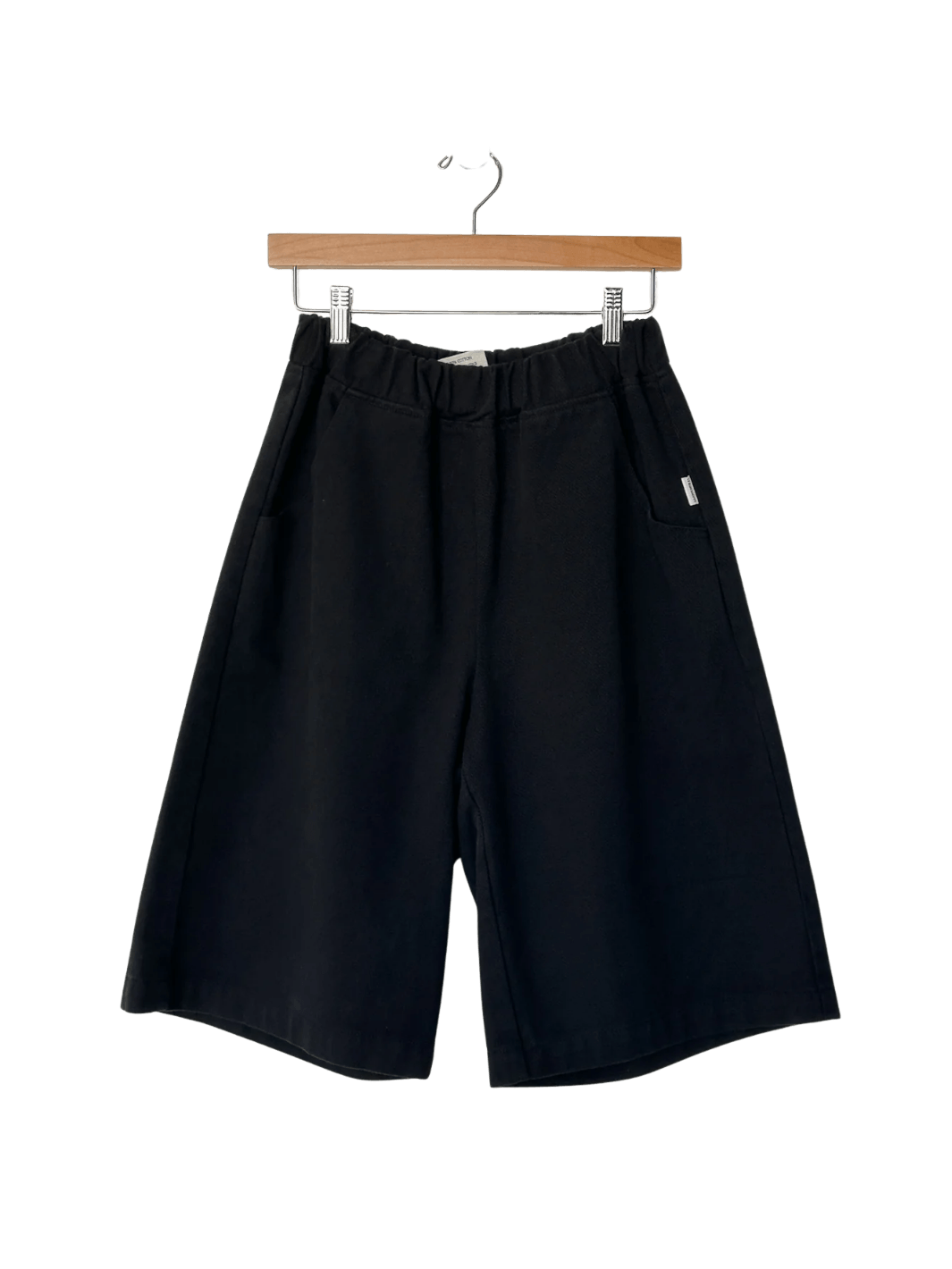 The Le Bon Shoppe Liam Bermuda Shorts - Black Canvas are displayed hanging from a wooden clip hanger | Twentyseven Toronto