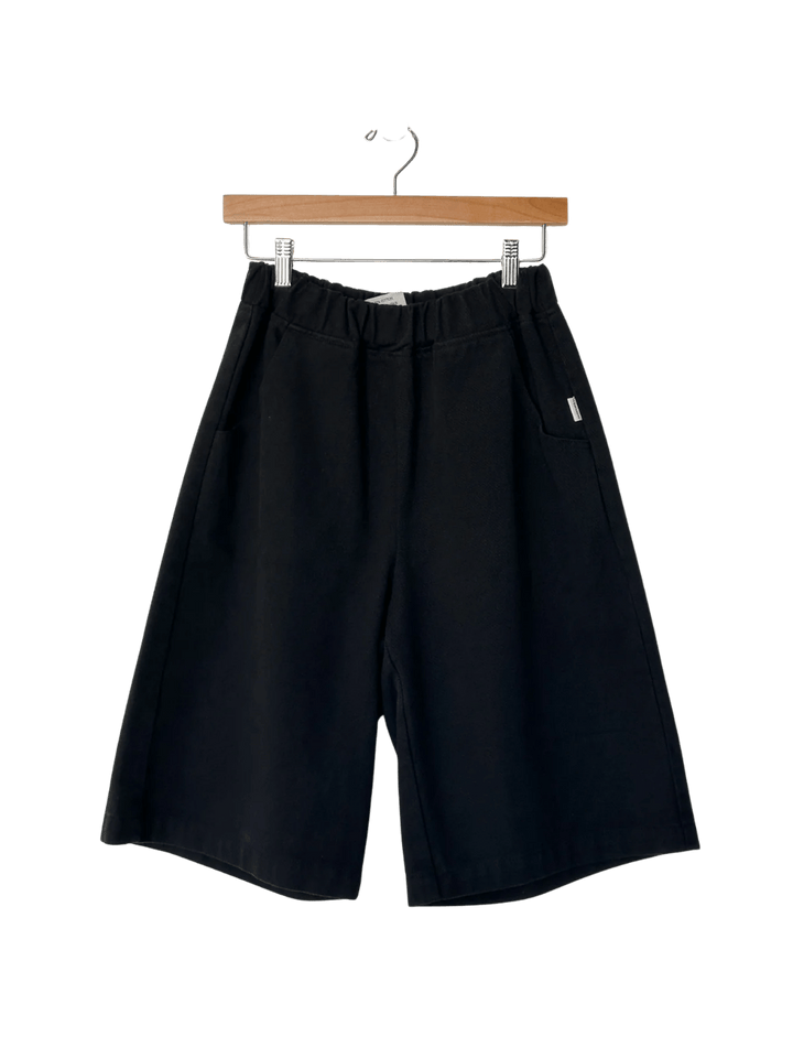 The Le Bon Shoppe Liam Bermuda Shorts - Black Canvas are displayed hanging from a wooden clip hanger | Twentyseven Toronto