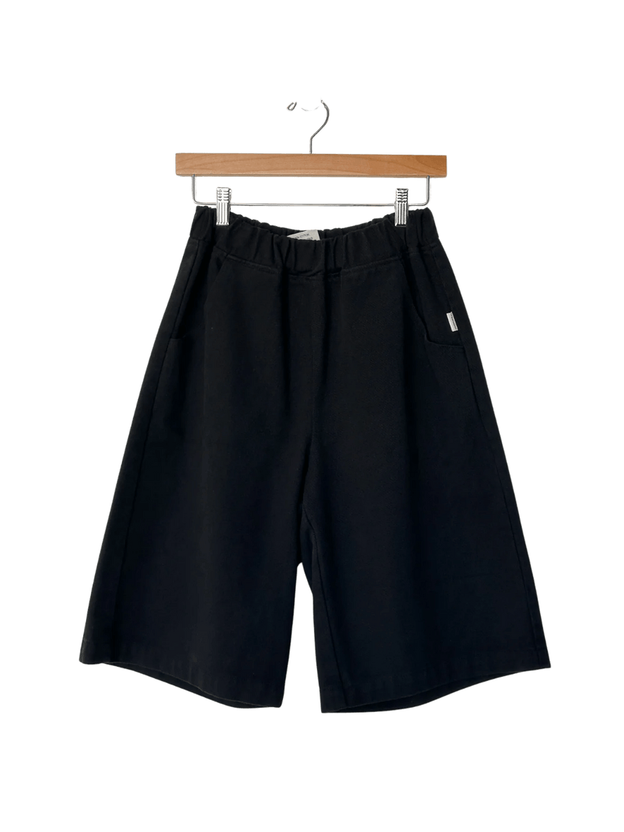 The Le Bon Shoppe Liam Bermuda Shorts - Black Canvas are displayed hanging from a wooden clip hanger | Twentyseven Toronto