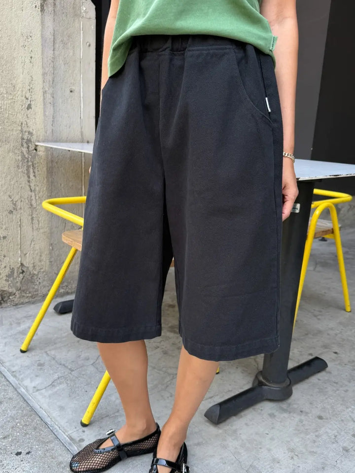 A person modeling the Le Bon Shoppe Liam Bermuda Shorts - Black Canvas next to an outdoor cafe table, highlighting the deep side pockets and relaxed fit | Twentyseven Toronto