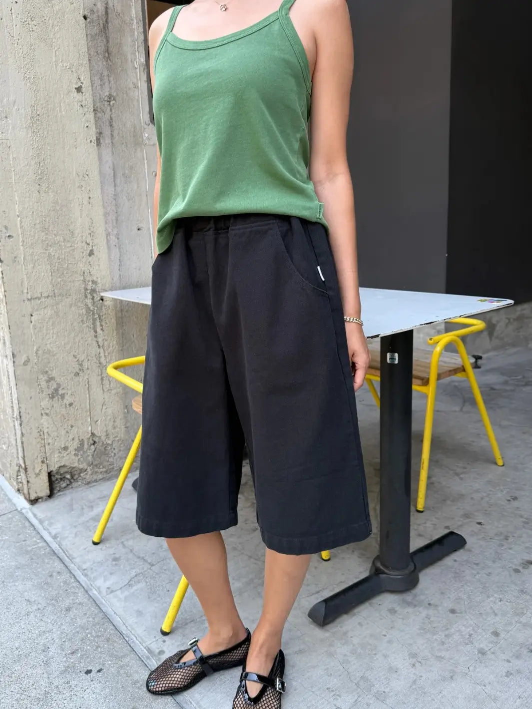 A person stands outside by a bistro table showcasing the elastic waistband and structured silhouette of the Le Bon Shoppe Liam Bermuda Shorts - Black Canvas paired with a green tank top and mesh flats | Twentyseven Toronto