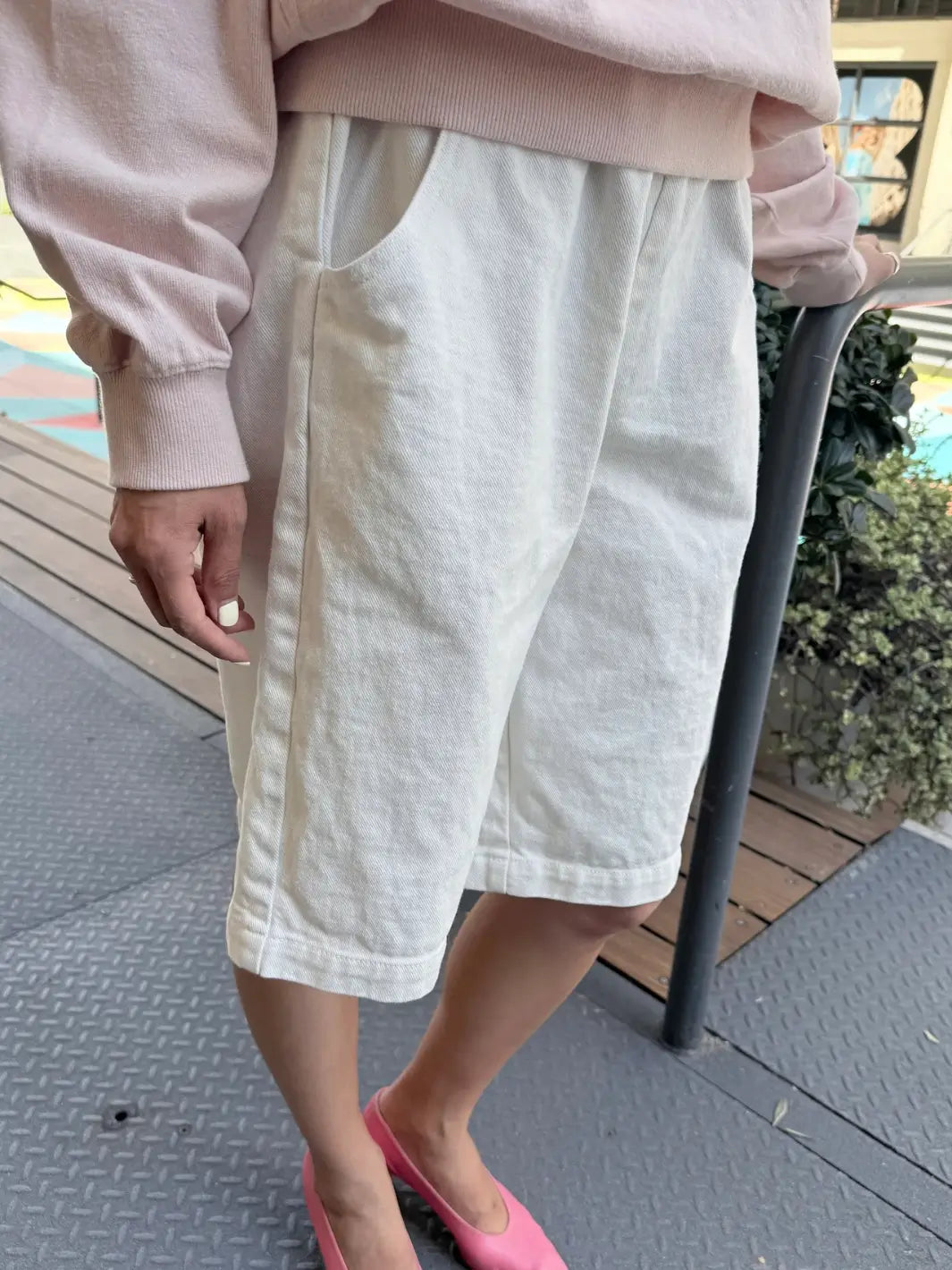 A close-up, side-angle view shows a person modeling the off-white Le Bon Shoppe Liam Bermuda Shorts - Milk paired with a light pink sweatshirt and pink heels | Twentyseven Toronto