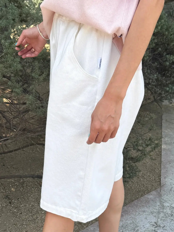 A side profile view of a person wearing the off-white Le Bon Shoppe Liam Bermuda Shorts - Milk, showcasing the deep side pocket and a small brand logo tag | Twentyseven Toronto