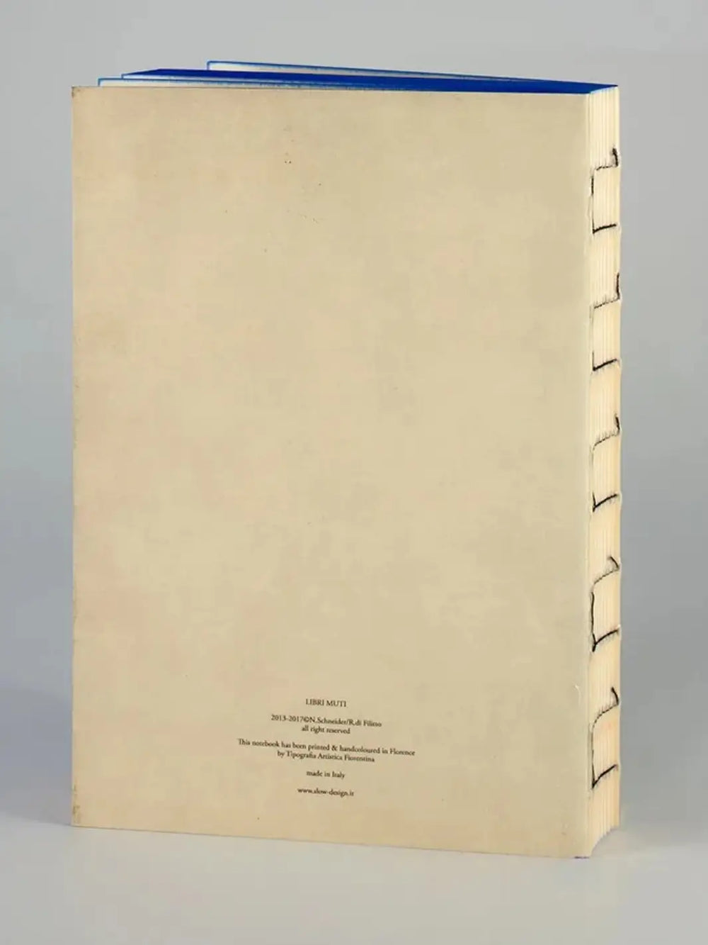 The back cover of a Moby Dick inspired notebook from Slow Design Libri Muti collection showing a clean, cream-colored surface with small publishing details at the bottom, an exposed hand-stitched spine, and blue hand-painted edges along the top | Twentyseven Toronto