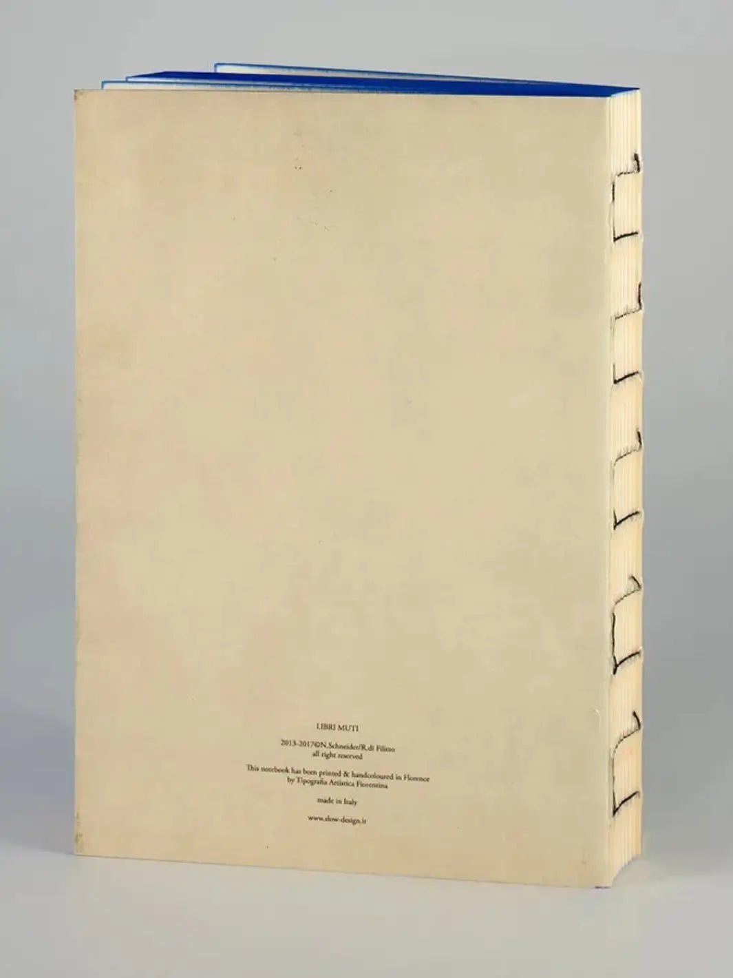 The back cover of a Moby Dick inspired notebook from Slow Design Libri Muti collection showing a clean, cream-colored surface with small publishing details at the bottom, an exposed hand-stitched spine, and blue hand-painted edges along the top | Twentyseven Toronto