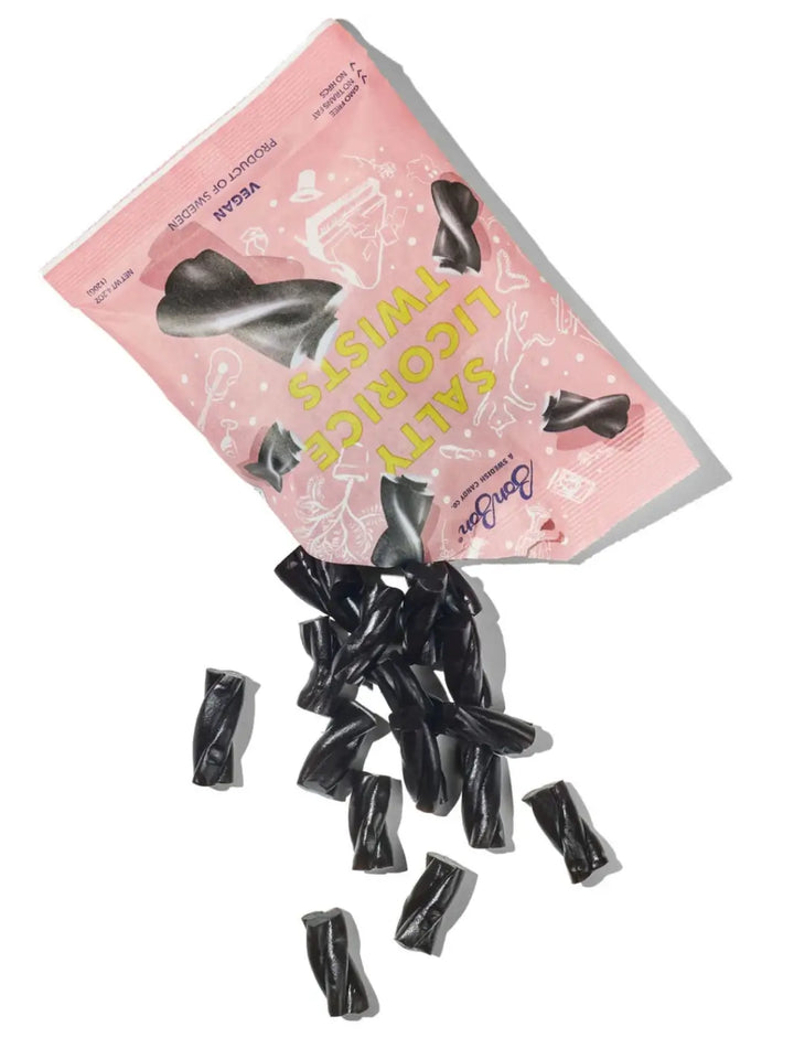 A handful of black, twisted licorice pieces spill out from an open pink pouch of Licorice Twist Swedish Candy from BonBon NYC onto a clean white surface | Twentyseven Toronto