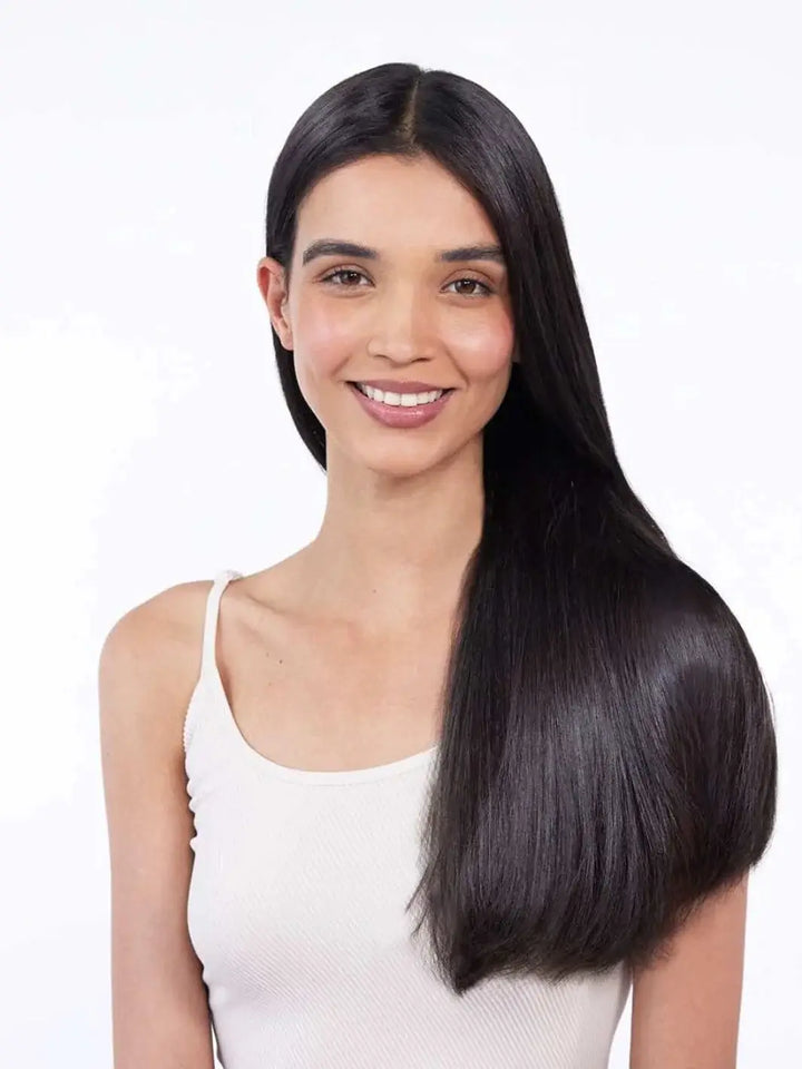 A smiling woman with exceptionally sleek, glossy straight dark hair draping over her shoulder demonstrates the smoothing effects of Hair by Sam McKnight Light Nourish Conditioner | Twentyseven Toronto