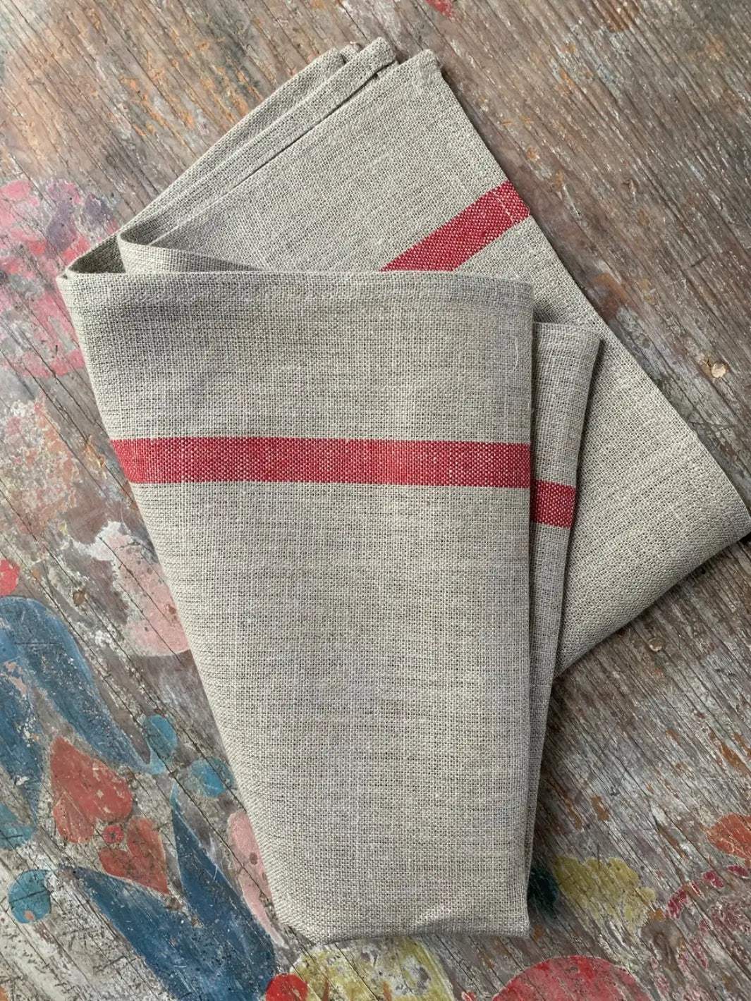 Folded Lined Kitchen Cloth - Natural with Red Stripe from Fog Linen Work rests on a distressed, colourful wooden tabletop showcasing its textured weave | Twentyseven Toronto