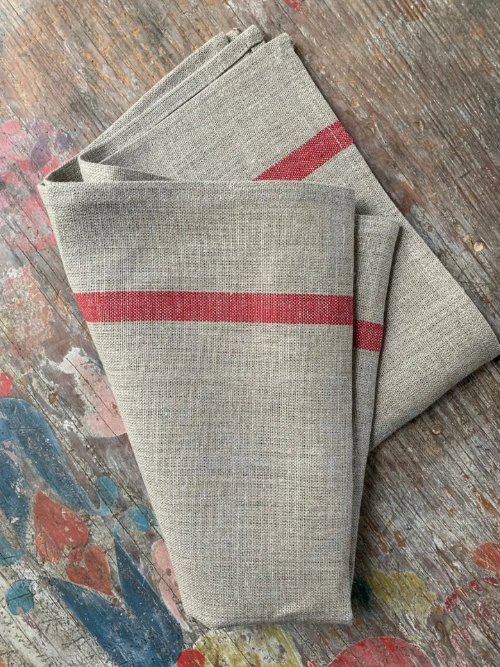 Folded Lined Kitchen Cloth - Natural with Red Stripe from Fog Linen Work rests on a distressed, colourful wooden tabletop showcasing its textured weave | Twentyseven Toronto