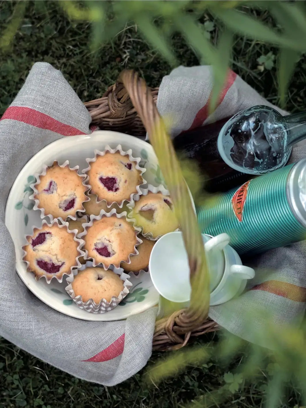 A woven picnic basket is lined with a Lined Kitchen Cloth - Natural with Red Stripe from Fog Linen Work, holding a plate of raspberry muffins and drinks | Twentyseven Toronto