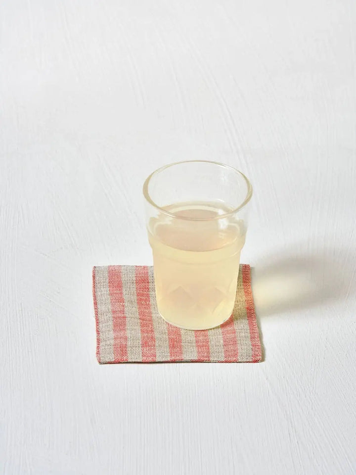 A clear glass of juice sits on a red and natural striped Fog Linen Work Linen Coaster - Agnes against a white background | Twentyseven Toronto